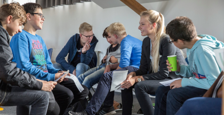 Junge Erwachsene sitzen zusammen mit Jugendlichen und unterhalten sich.