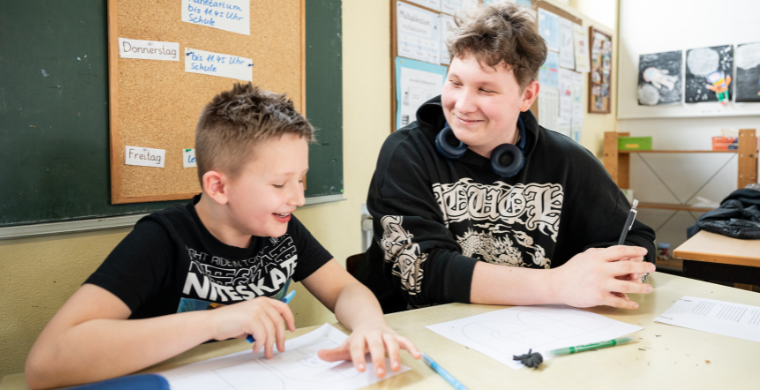 Ein Grundschulkind und ein Jugendlicher sitzen lachend nebeneinander an einem Tisch und arbeiten miteinander.