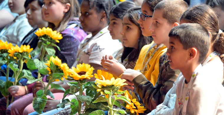 Grundschulkinder sitzen in einer Reihe nebeneinander und schauen gespannt in dieselbe Richtung. Sie halten Sonnenblumen in den Händen.