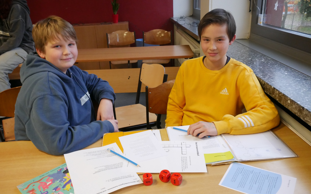 Zwei 14-jährige Jungs sitzen in einem Klassenzimmer nebeneinander am Tisch. Darauf ausgebreitet liegt Material das sie für ihr Arbeit als Coachs für Grundschulkinder nutzen können.